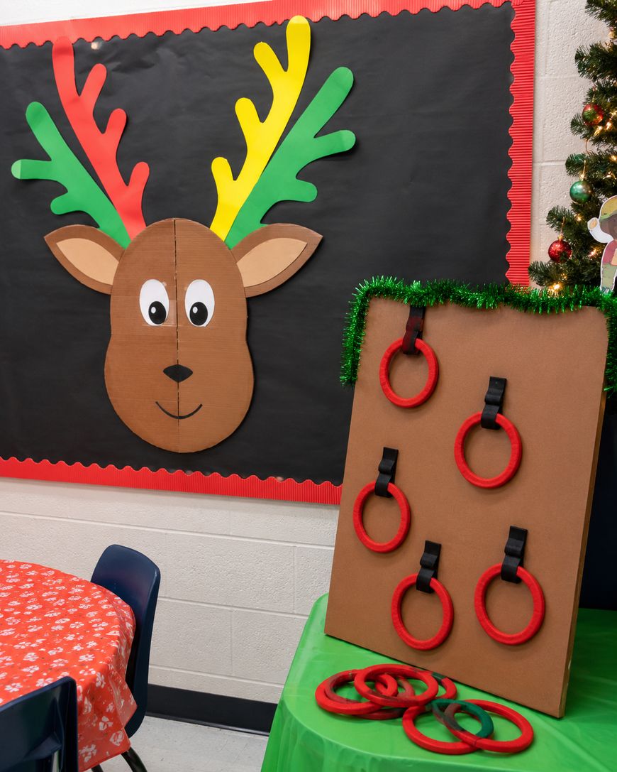 Classroom with a large reindeer face and antlers on the wall. In front, theres a ring toss game board with red and green rings. A table with a red cloth and blue chairs is nearby. Holiday decorations are present.