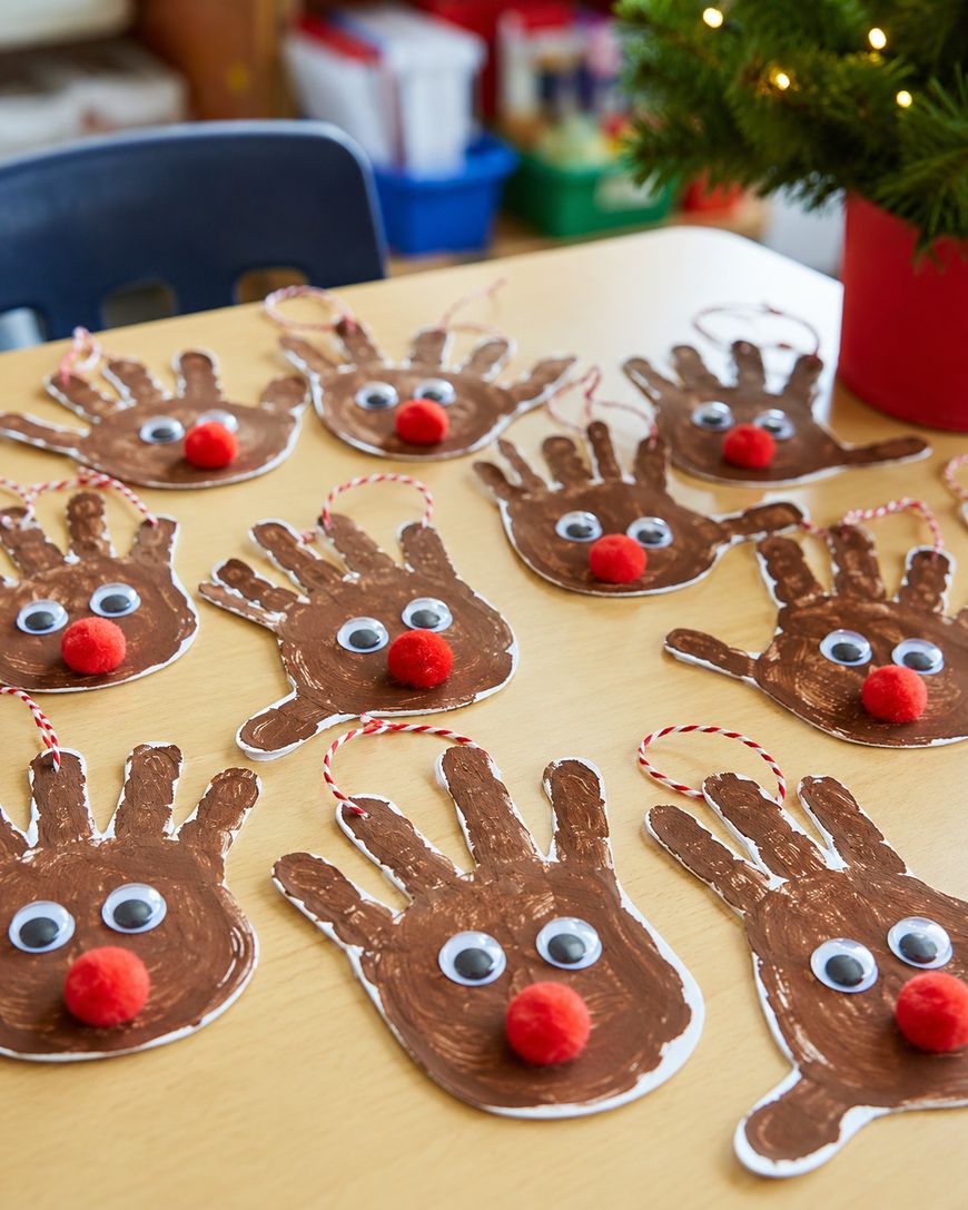 Several reindeer crafts made from brown painted handprints with googly eyes, red pom-pom noses, and string hangers are arranged on a table. A small decorated tree is partially visible in the corner.