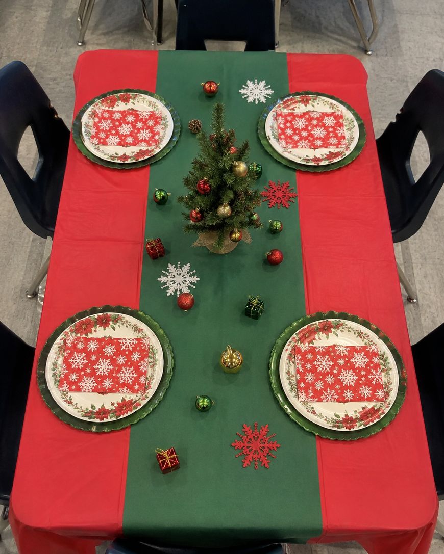 A festive table set for four with red and green decor, snowflake plates, napkins, small ornaments, and a mini Christmas tree centerpiece. Four black chairs surround the table.