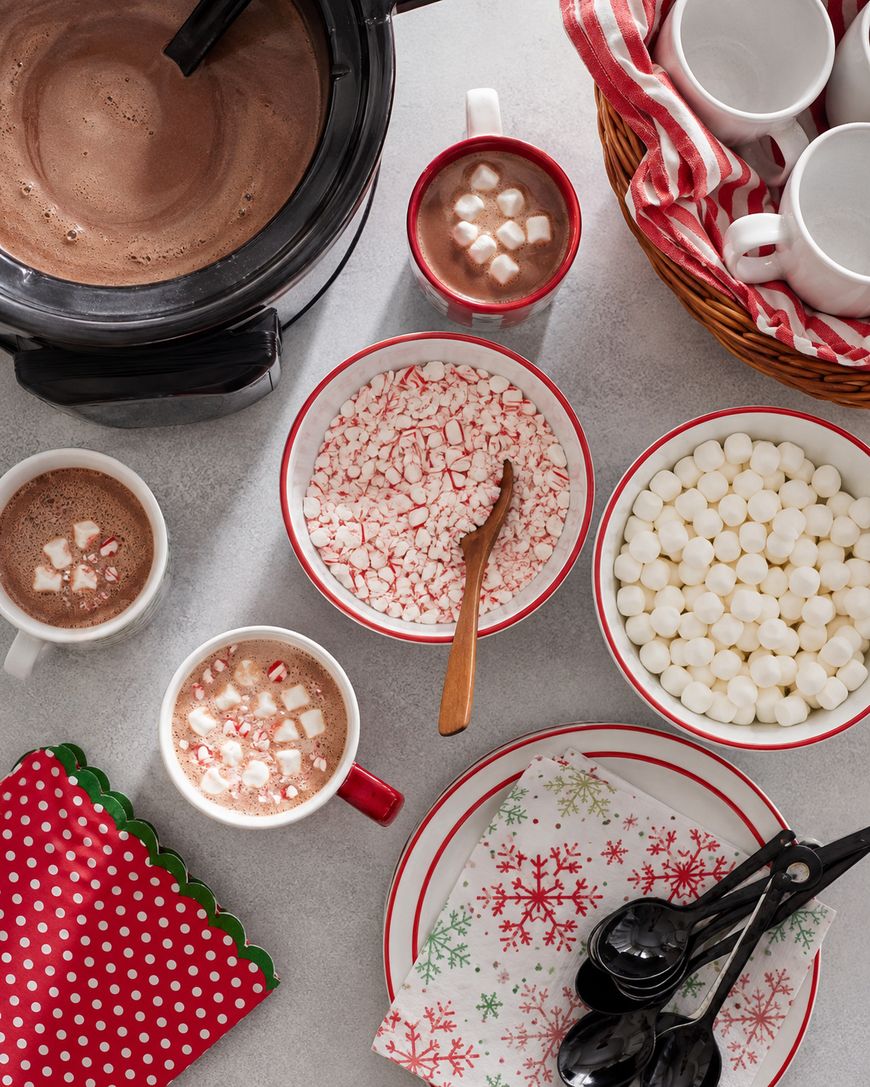A hot chocolate bar with mugs, a crockpot of hot cocoa, bowls of mini marshmallows and crushed peppermint, festive napkins, black spoons, and a basket of stacked white mugs with candy-cane striped cloth.