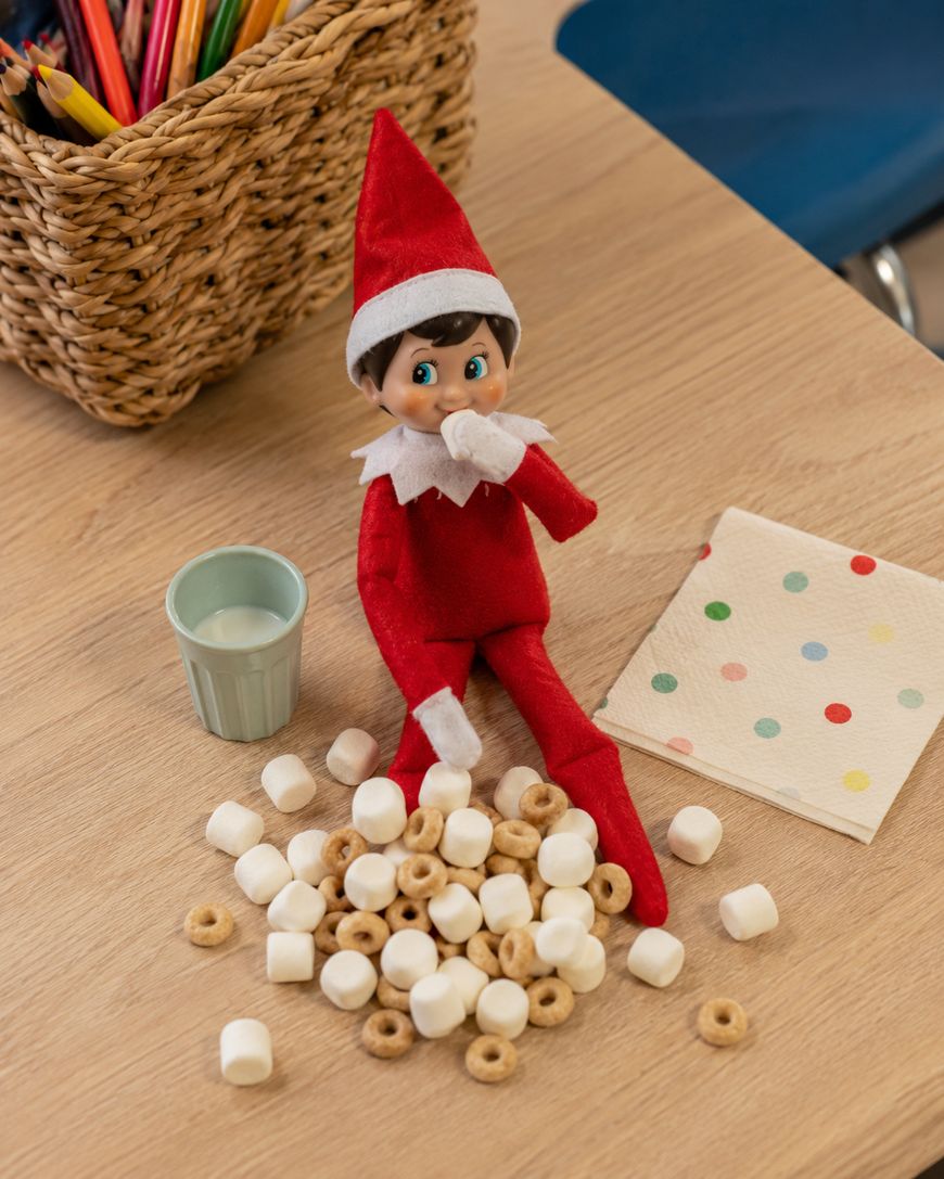 A toy elf in red clothes sits on a wooden table, surrounded by marshmallows and cereal. Theres a small cup, a napkin with colorful dots, and a basket of colored pencils nearby.