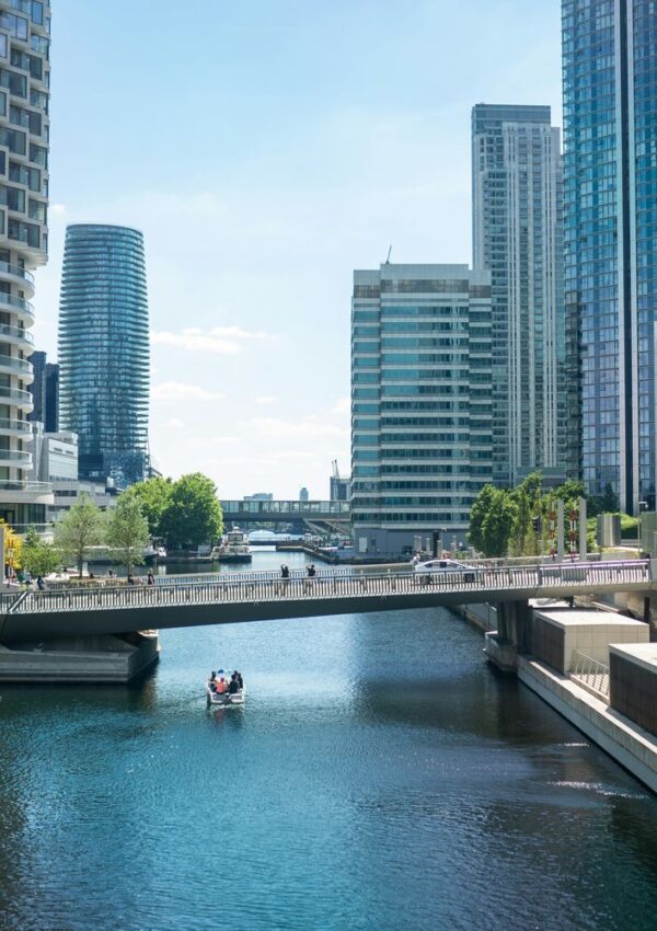 A group of people ride a small boat on a canal bordered by modern high-rise buildings under a blue sky, with a pedestrian bridge crossing above the water.