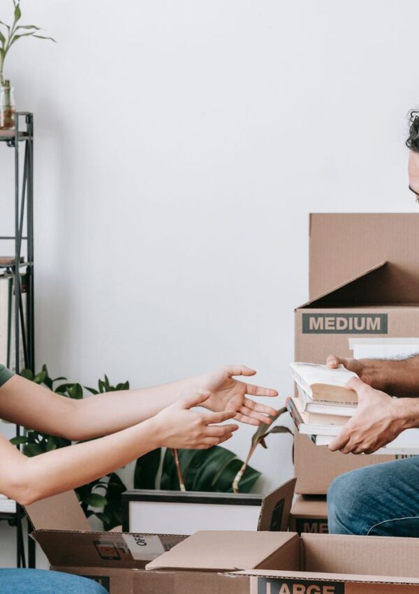 Two people sit near cardboard boxes, passing a stack of books between them. There is a shelf with plants and books in the background, and the scene suggests packing or unpacking in a home setting.