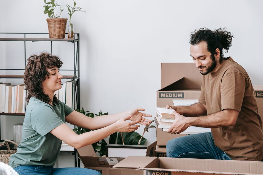 Two people packing books into cardboard boxes, smiling and working together in a bright room with plants and shelves in the background.
