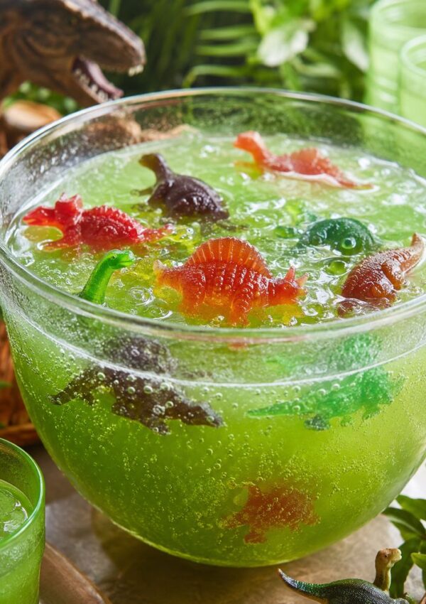 A large glass bowl of bright green punch with gummy dinosaurs floating in it, surrounded by greenery and dinosaur decorations, creating a playful, prehistoric theme.
