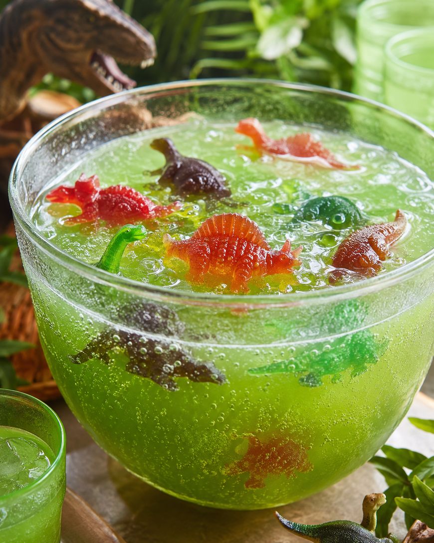 A large glass bowl filled with bright green fizzy punch, with colorful plastic dinosaur toys floating inside. There are green cups and leafy decorations in the background.
