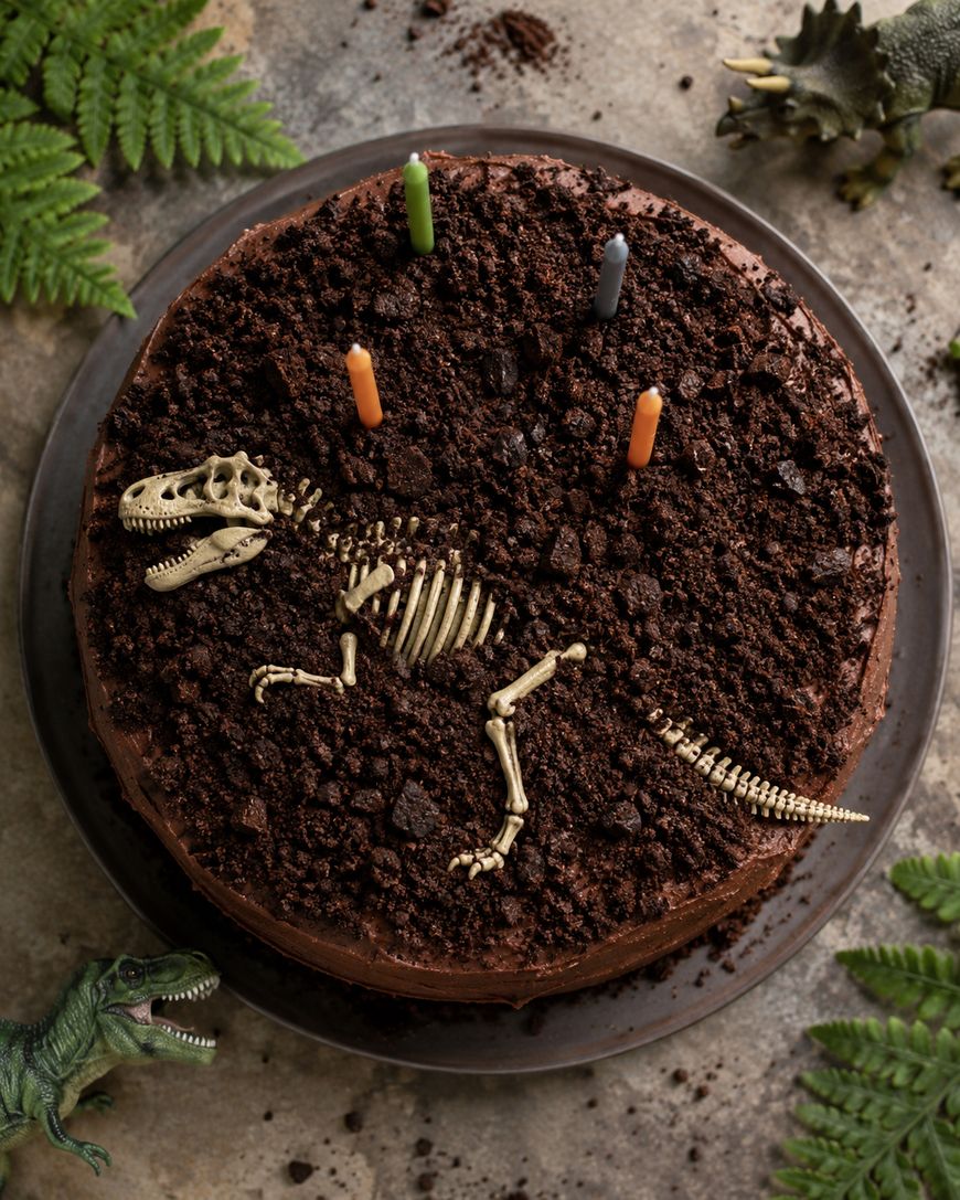 A chocolate cake topped with crumbled cookies, a plastic dinosaur skeleton, and four colorful candles, surrounded by toy dinosaurs and fern leaves.