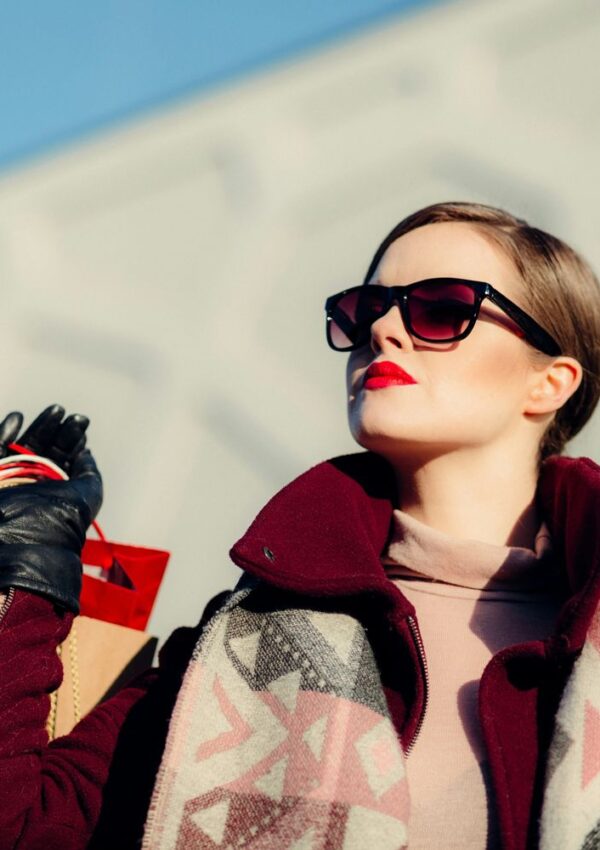A stylish woman wearing sunglasses, red lipstick, and a patterned scarf holds shopping bags outdoors on a sunny day, effortlessly showcasing how simple style swaps can elevate everyday looks.