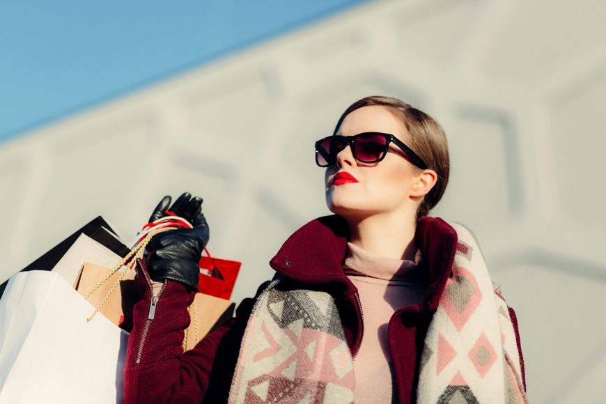 A stylish woman in sunglasses and red lipstick holds several shopping bags outdoors, demonstrating how Simple Style Swaps—like her patterned scarf and maroon coat—can elevate Everyday Looks against a modern light gray background.