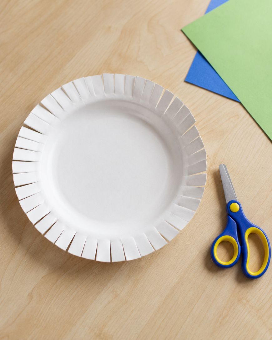 A white paper plate with evenly cut slits around the edge sits on a wooden table next to blue and green paper sheets and a pair of scissors with blue and yellow handles.