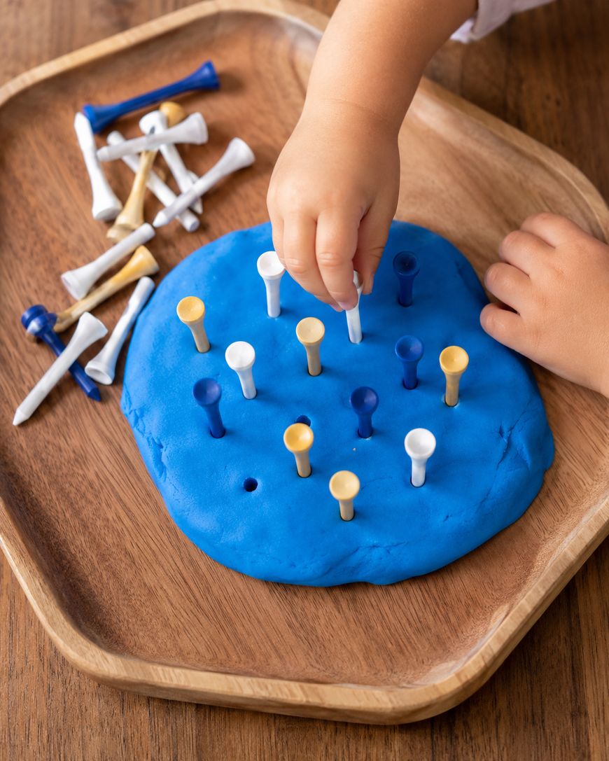 A childs hand places a golf tee into blue playdough on a wooden tray, with extra golf tees in the background.