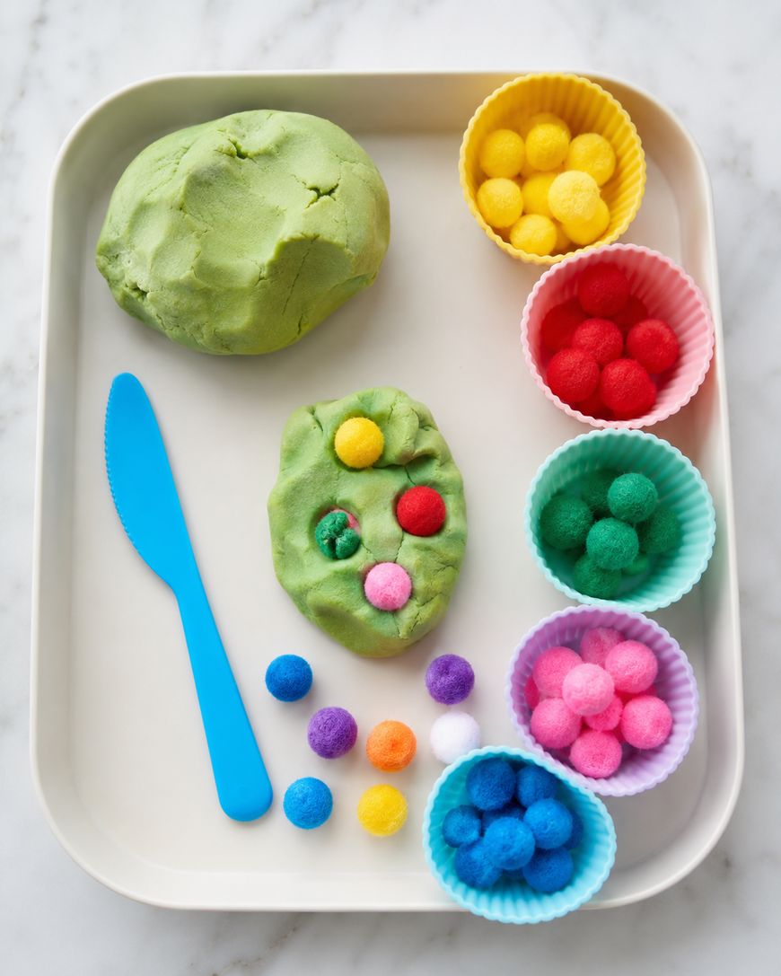 A tray with green playdough, a blue plastic knife, and colorful pom-poms in silicone cups. Some pom-poms are pressed into the playdough. The scene is set on a white marble surface.