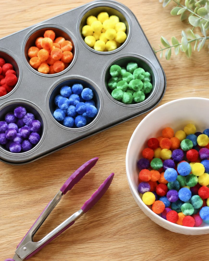 A muffin tin filled with colorful pom-poms, sorted by color, sits on a wooden table next to a bowl of mixed pom-poms, metal tongs with purple tips, and a green leafy plant.