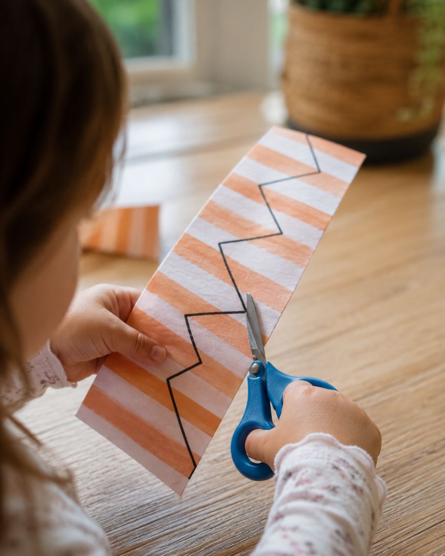 A child uses blue scissors to cut along a zigzag line drawn on a striped piece of paper.