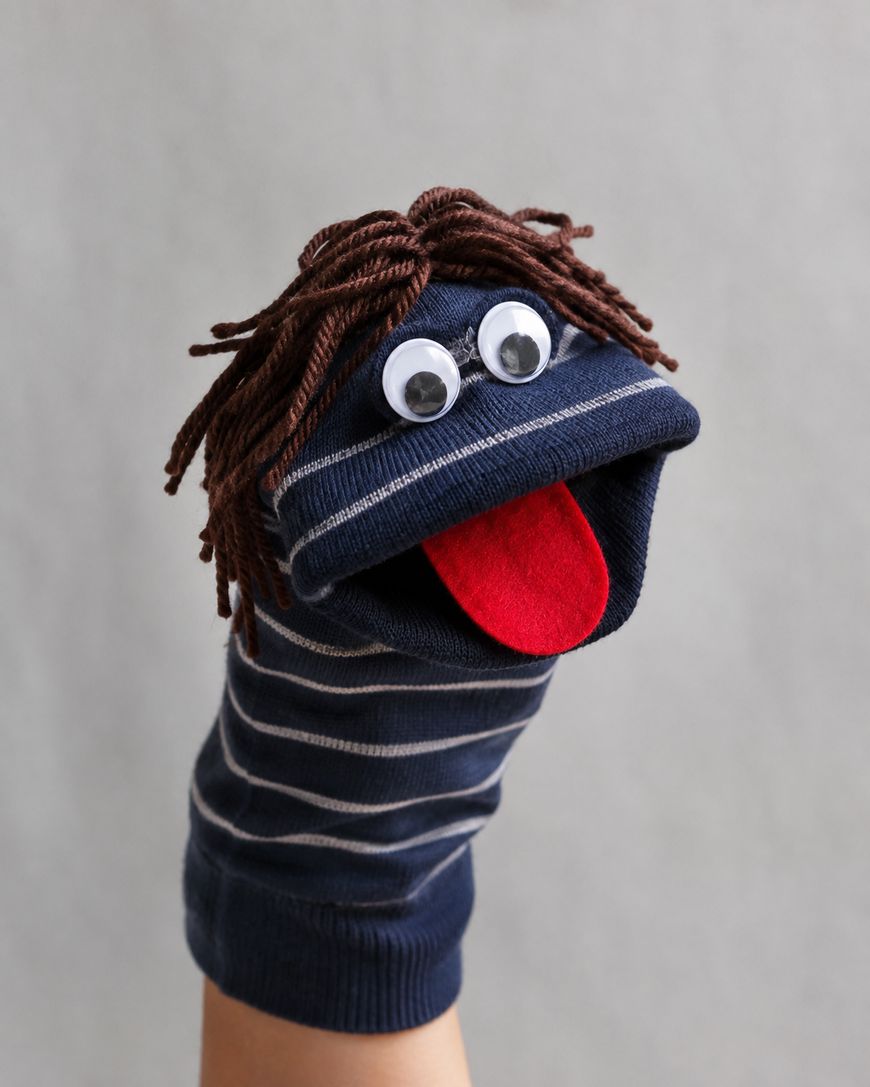 A striped navy blue sock puppet with googly eyes, brown yarn hair, and a red felt tongue is worn on a hand against a plain gray background.