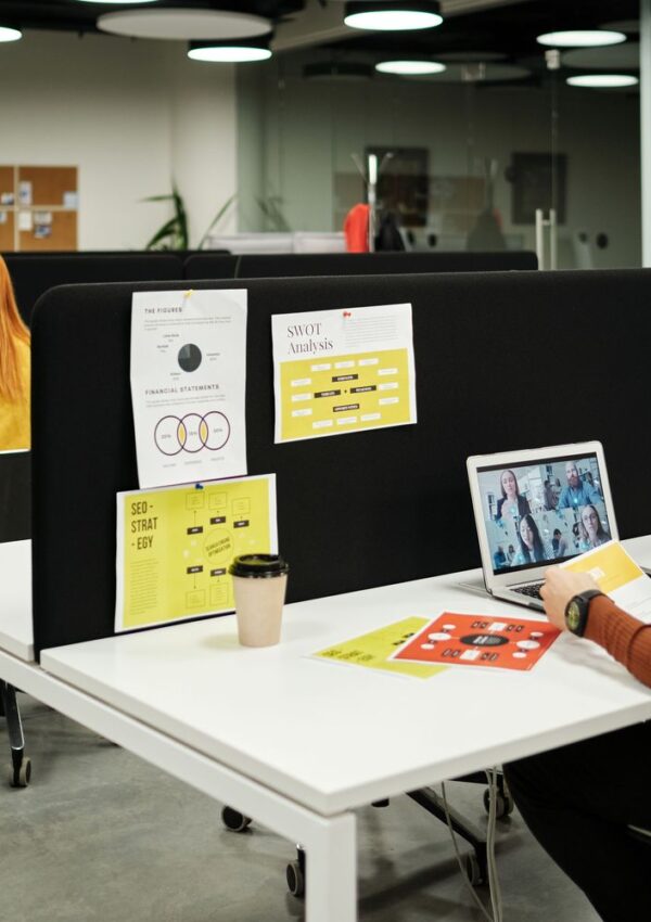 Office workspace with laptops, papers, coffee cup, and printed charts on a partition. One person works on a video call while another sits across, both partially visible. The setting reflects a healthier office environment, with background elements like glass walls and a corkboard supporting collaboration and office wellness.
