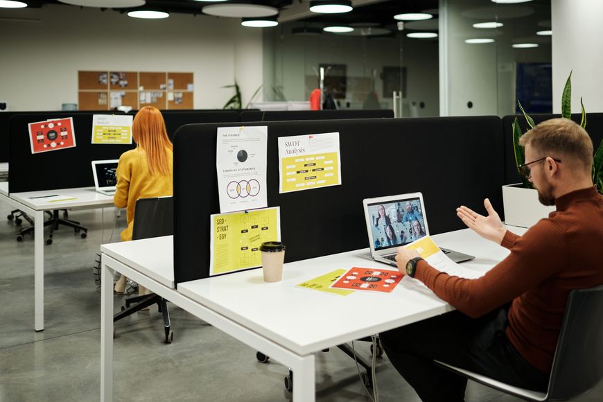 Two people work at desks separated by partitions in a modern office designed to promote office wellness. One man gestures at his laptop during a video call, while a woman works on a computer. Papers and a coffee cup are on the desks, reflecting an environment focused on productivity and a healthier office environment.