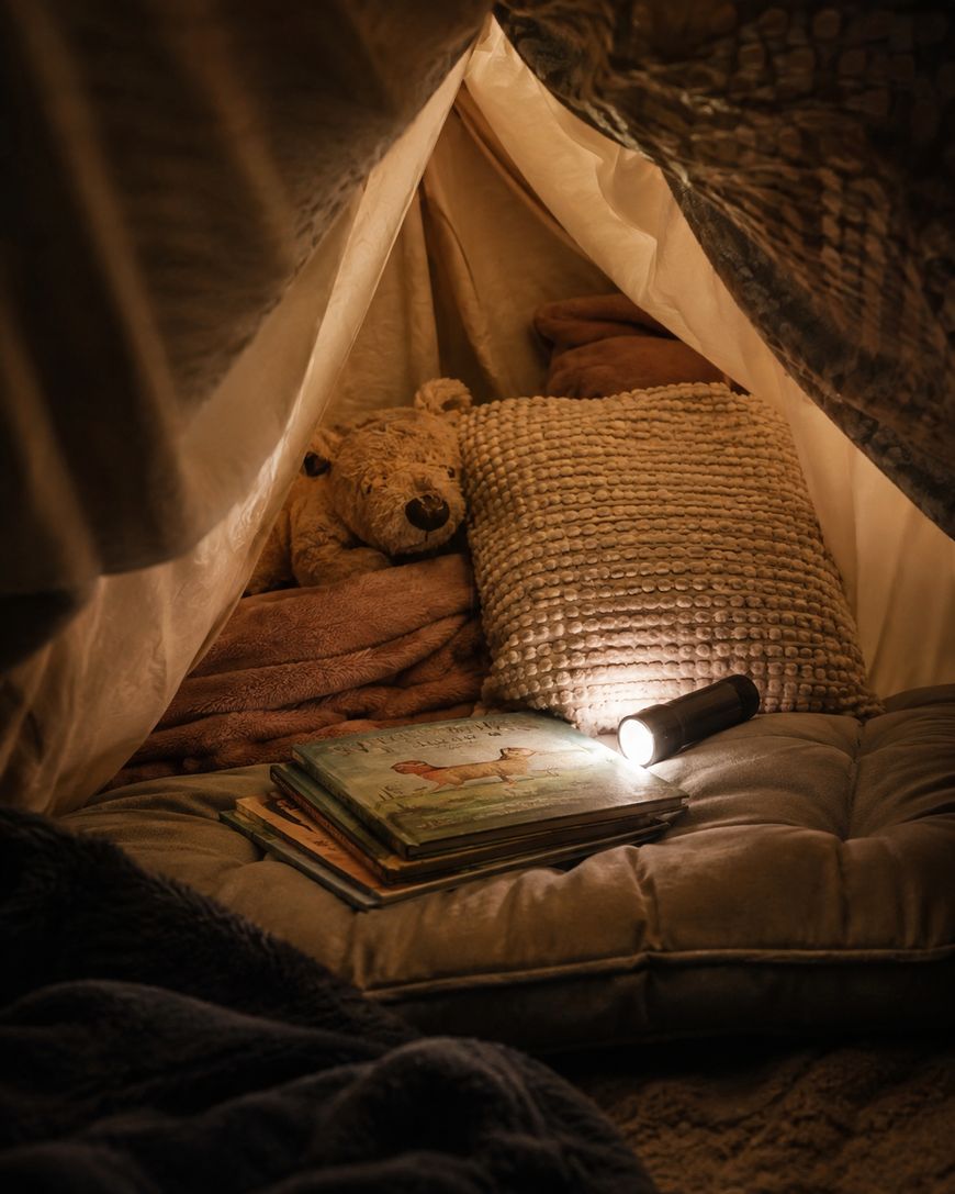 A cozy blanket fort with cushions, a stuffed bear, a pillow, a flashlight, and a stack of books, softly lit by warm light.