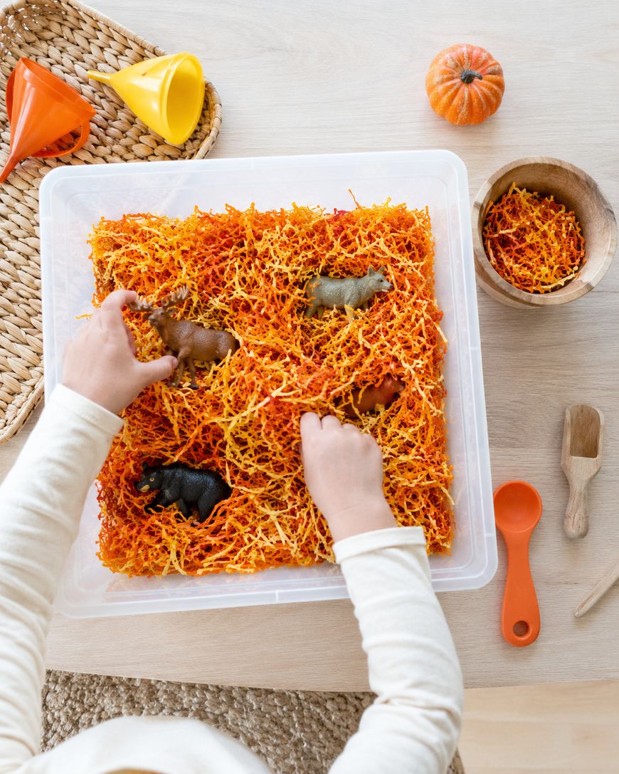 Childs hands playing with toy animals in a sensory bin filled with orange and yellow shredded paper. Nearby are a small pumpkin, wooden bowl, spoon, brush.