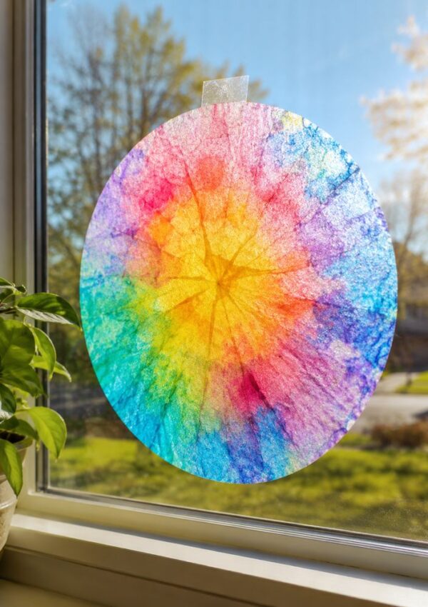 A colorful, circular suncatcher made from a coffee filter hangs on a sunny window with trees and grass outside. A potted plant sits on the windowsill.