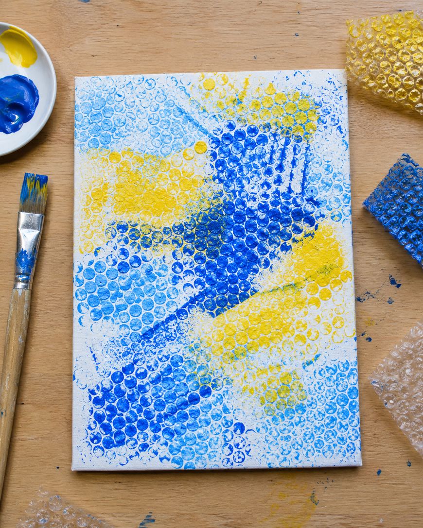 A canvas with blue and yellow bubble wrap paint prints, a paintbrush with blue paint, a palette with blue and yellow paint, and pieces of used bubble wrap on a wooden table.