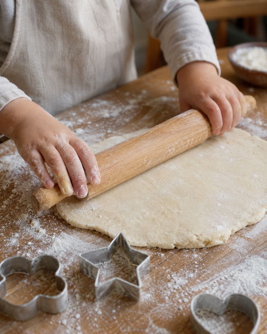 A child rolls out dough with a wooden rolling pin on a floured surface. Metal cookie cutters in heart and star shapes are nearby.