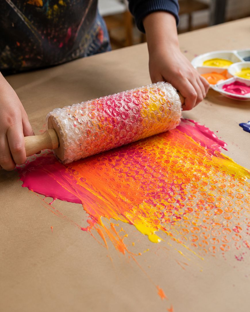 A person rolls a rolling pin covered in bubble wrap over red, orange, and yellow paint on brown paper, creating a textured pattern.