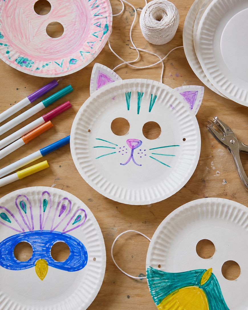 Colorful animal masks made from paper plates are shown on a wooden table with markers, string, a hole punch, and decorated plates resembling a cat, a bird, and a peacock.