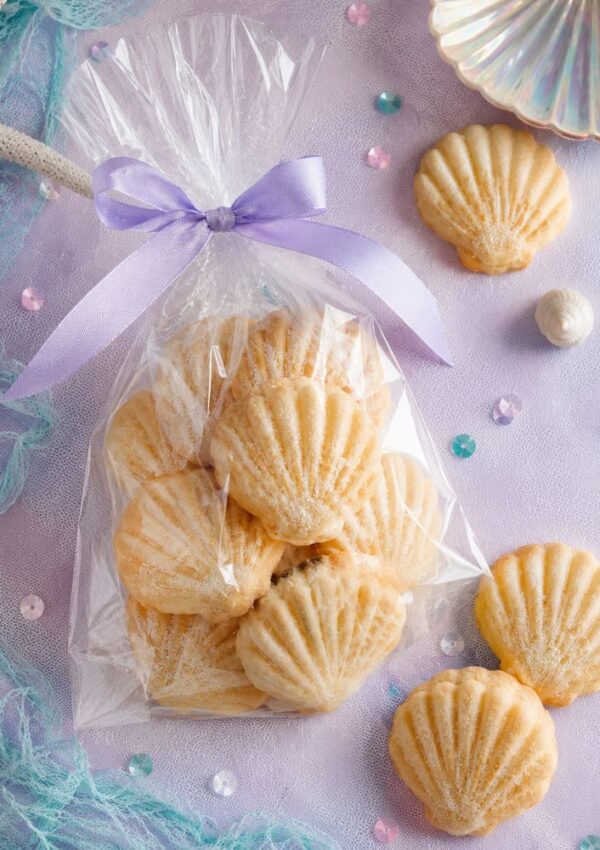A clear bag tied with a lavender ribbon holds shell-shaped cookies on a pastel purple cloth decorated with sequins, netting, and seashells.