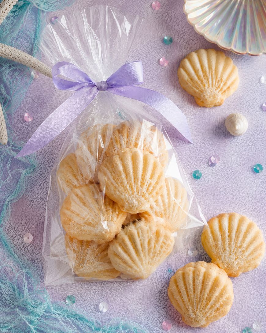 A clear bag tied with a lavender ribbon holds shell-shaped shortbread cookies, surrounded by loose shell cookies, pearls, and coral on a lavender surface.