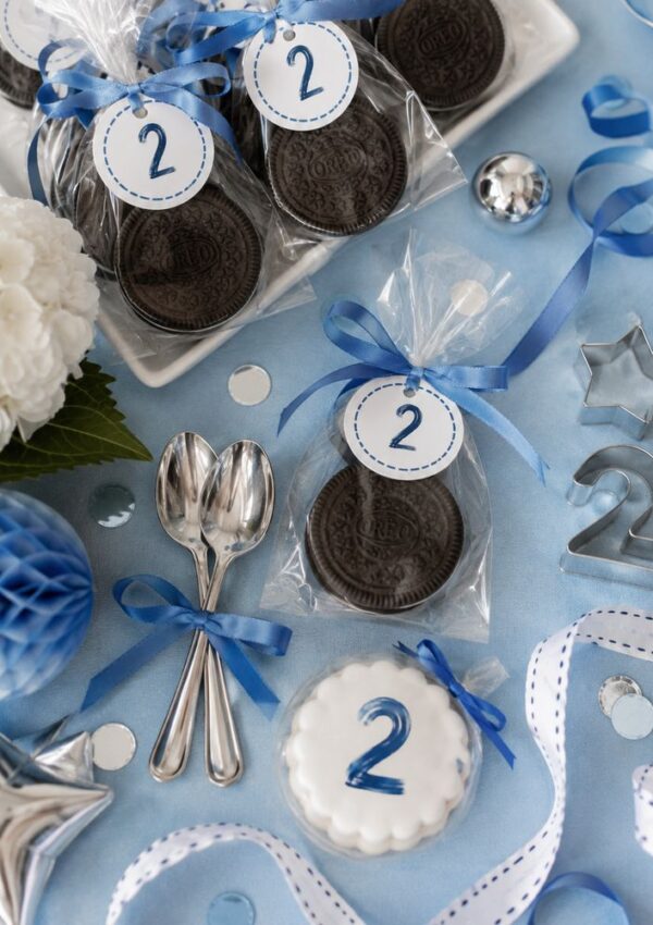 Blue-themed party table with Oreos in bags labeled 2, star and number cookie cutters, silver spoons tied with blue ribbon, a white cookie with a blue 2, flowers, confetti, and blue ribbon decorations.
