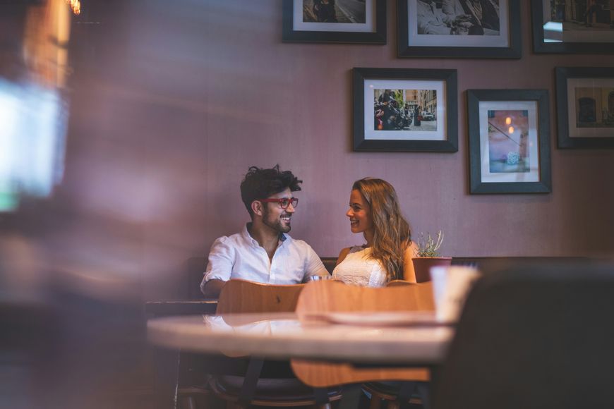 A man and woman sit smiling at each other in a cozy cafe, with framed photos on the wall behind them and a blurred foreground, creating a warm, intimate atmosphere.