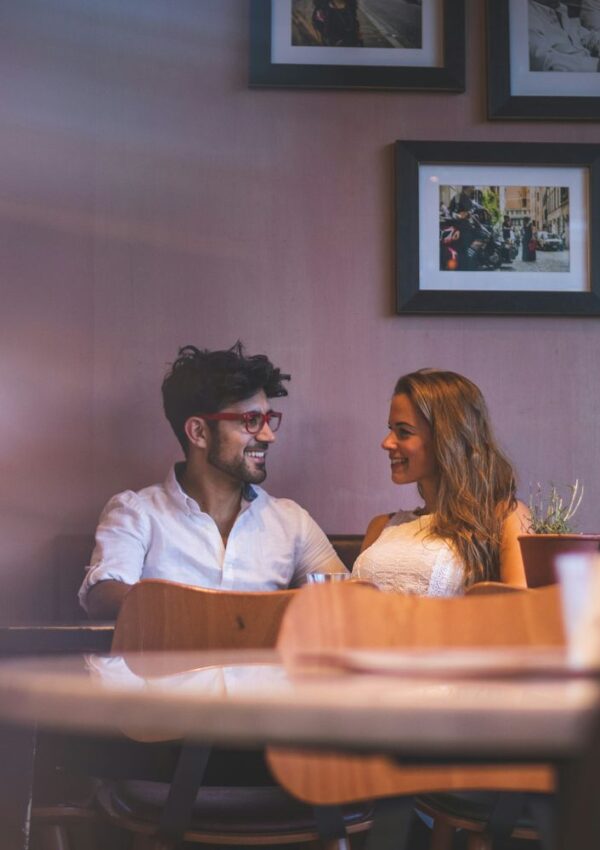 A man and woman sit together at a table in a cozy café, smiling at each other. Framed photos hang on the wall behind them, and soft light creates a warm, inviting atmosphere.