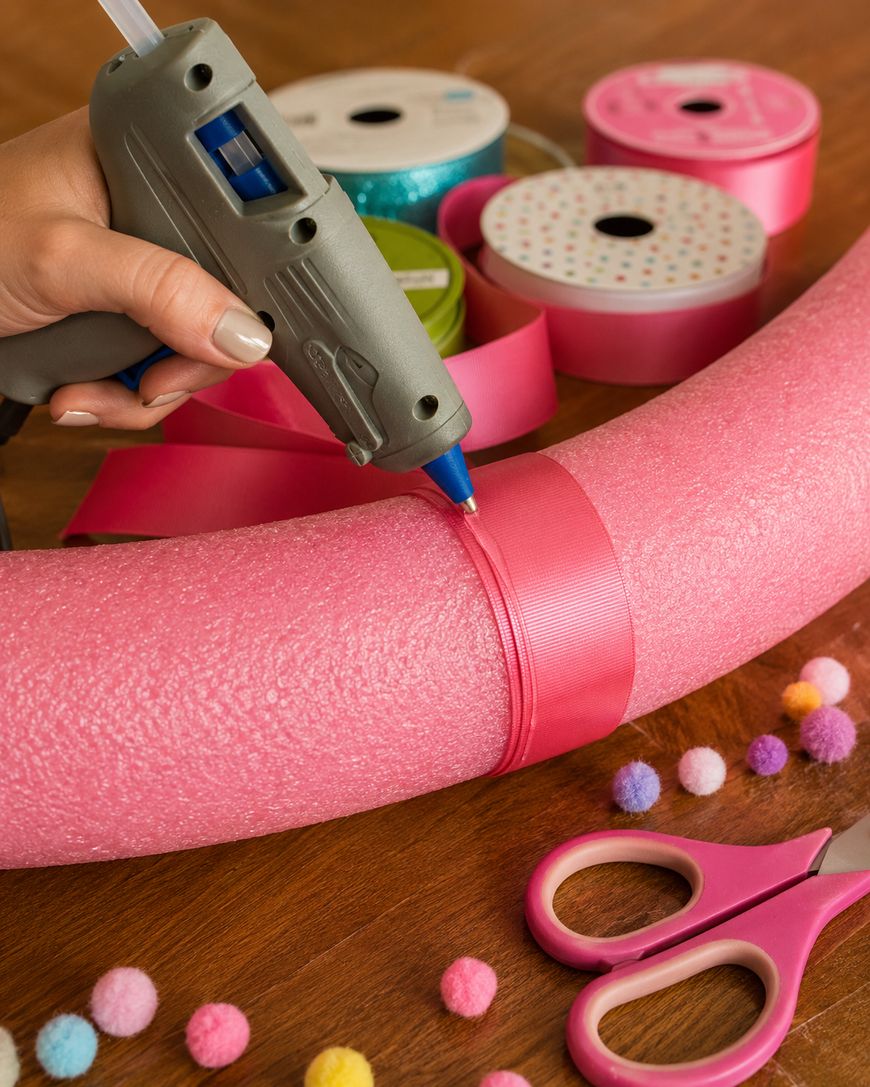 A hand uses a glue gun to attach a pink ribbon to a pink foam pool noodle. Scissors, ribbon spools, and colorful pom-poms are on the wooden table.