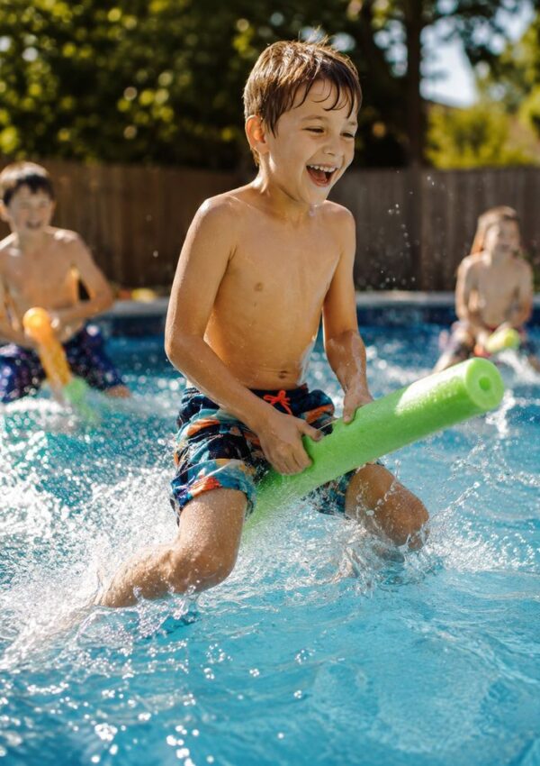 Three kids play in a pool with water noodles on a sunny day, splashing and laughing. The boy in front is smiling widely, wearing swim trunks, and sunlight shines through the water.