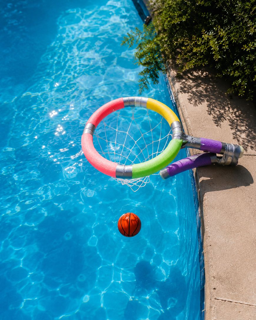 A colorful poolside basketball hoop is attached to the pool edge as an orange basketball heads toward the net over bright blue water. Green shrubs and concrete are visible beside the pool.