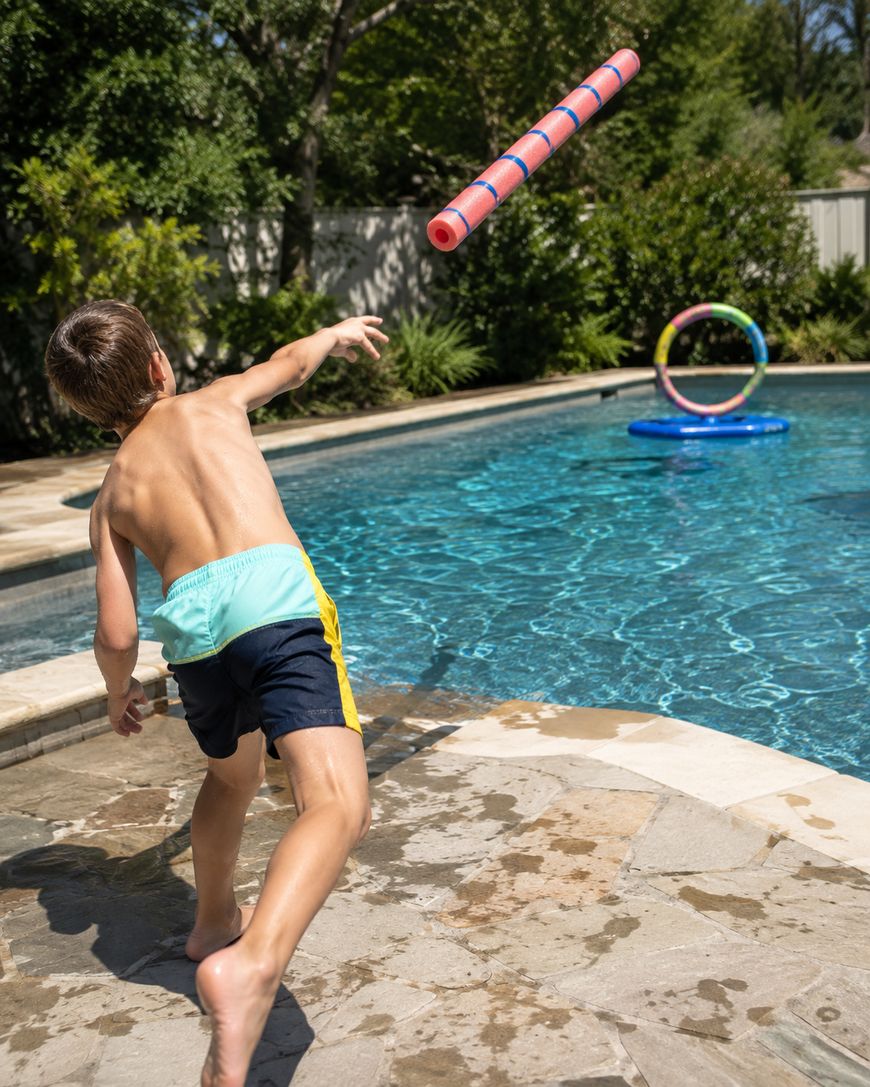 A boy in swim trunks throws a red pool noodle toward a floating ring target in a backyard swimming pool on a sunny day.