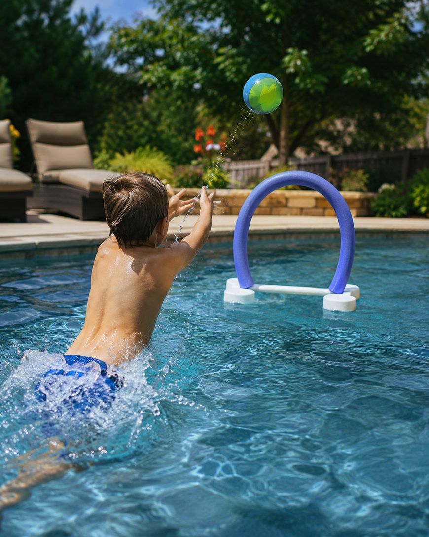 A boy in blue swim trunks throws a ball through a floating hoop in a backyard swimming pool on a sunny day. Lounge chairs and green trees are visible in the background.