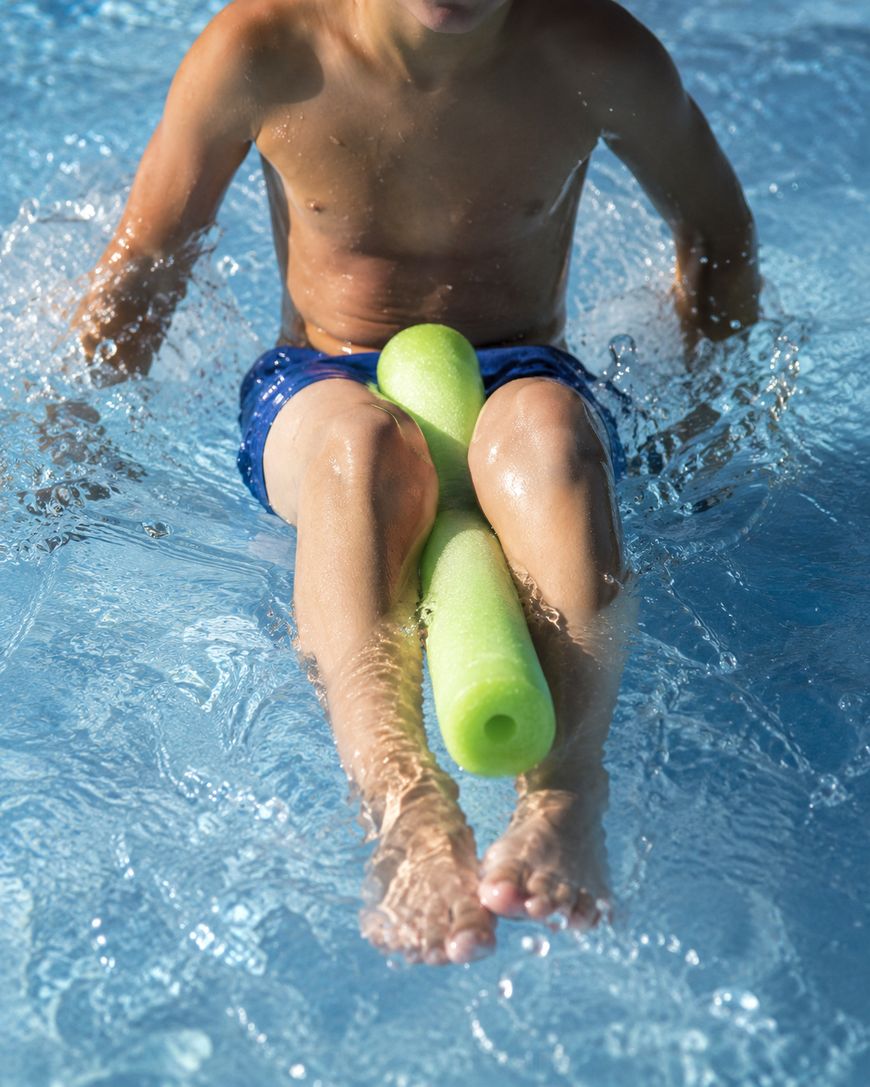 A child wearing blue swim trunks is sitting at the edge of a pool, splashing water and holding a green pool noodle between their legs.