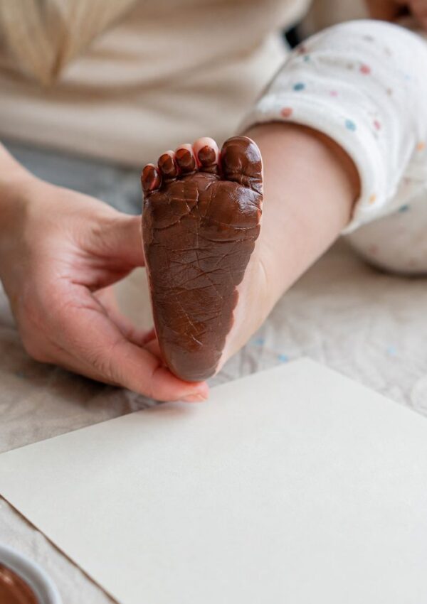 An adult holds a babys foot covered in brown paint, preparing to make a footprint on a blank sheet of paper.