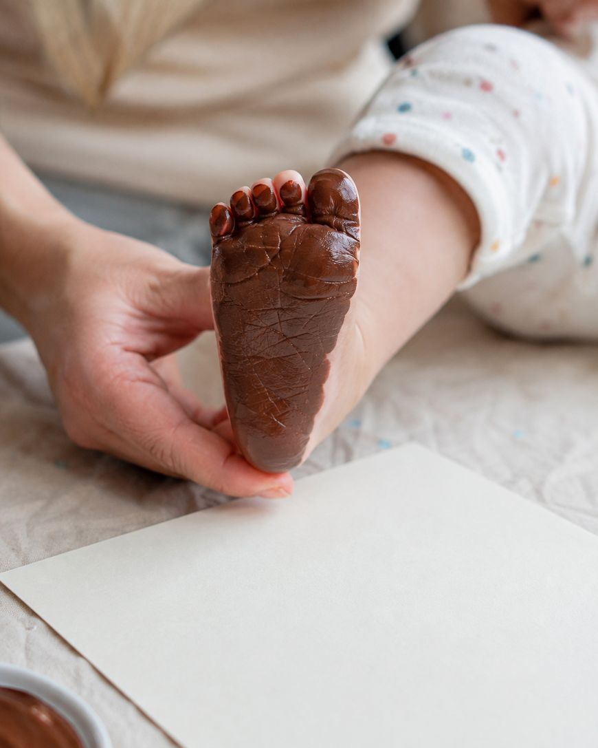 An adult holds a babys foot covered in brown paint, preparing to make a footprint on a blank sheet of paper.