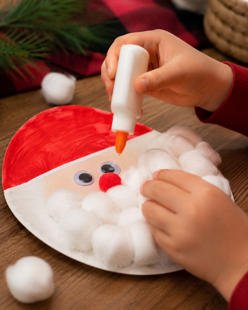 A child glues cotton balls onto a paper plate Santa craft with painted hat, googly eyes, and red nose. The scene includes holiday decor and a wooden table.