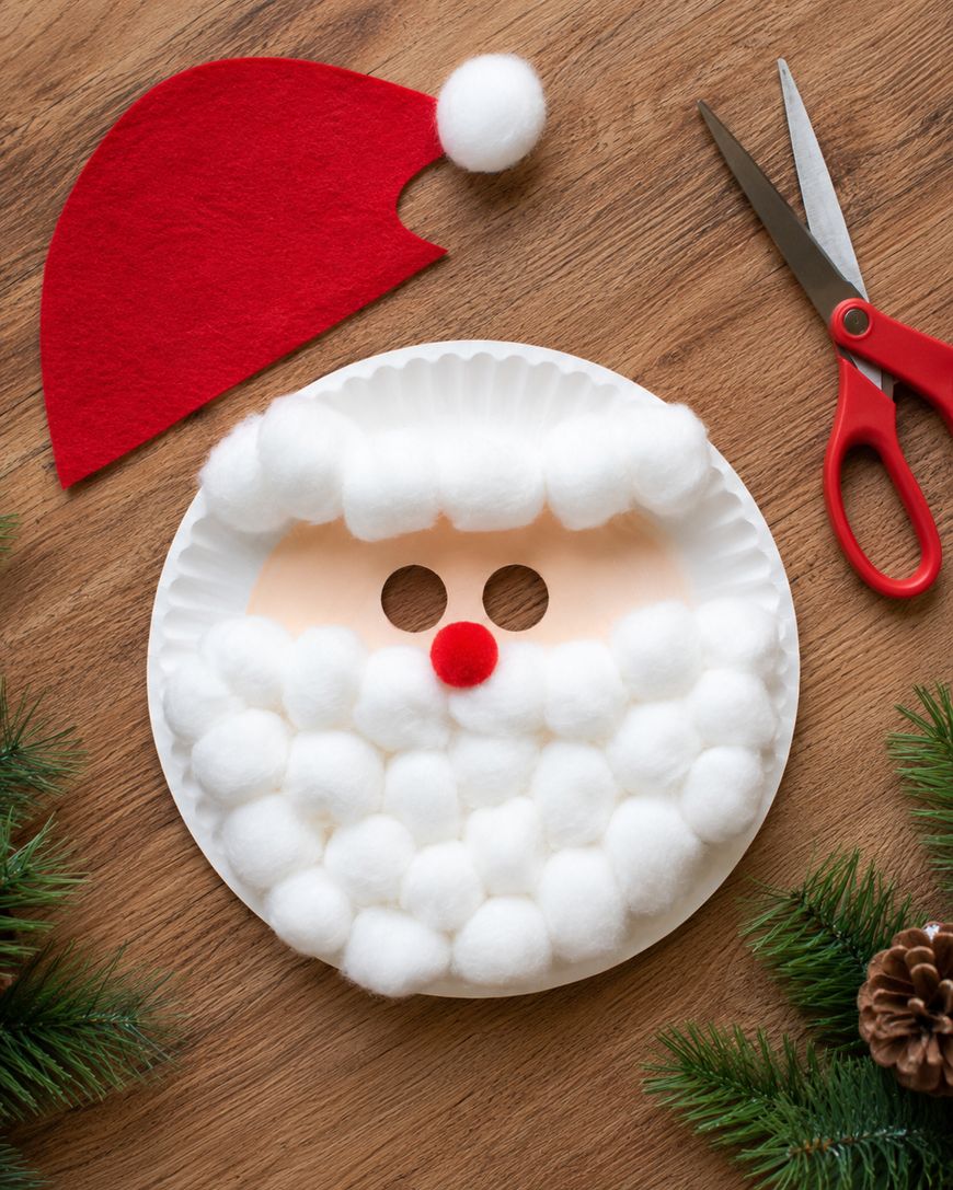 Paper plate Santa mask decorated with cotton balls and a red pompom nose, red felt hat nearby, red scissors, pinecones, and pine branches on a wooden table.