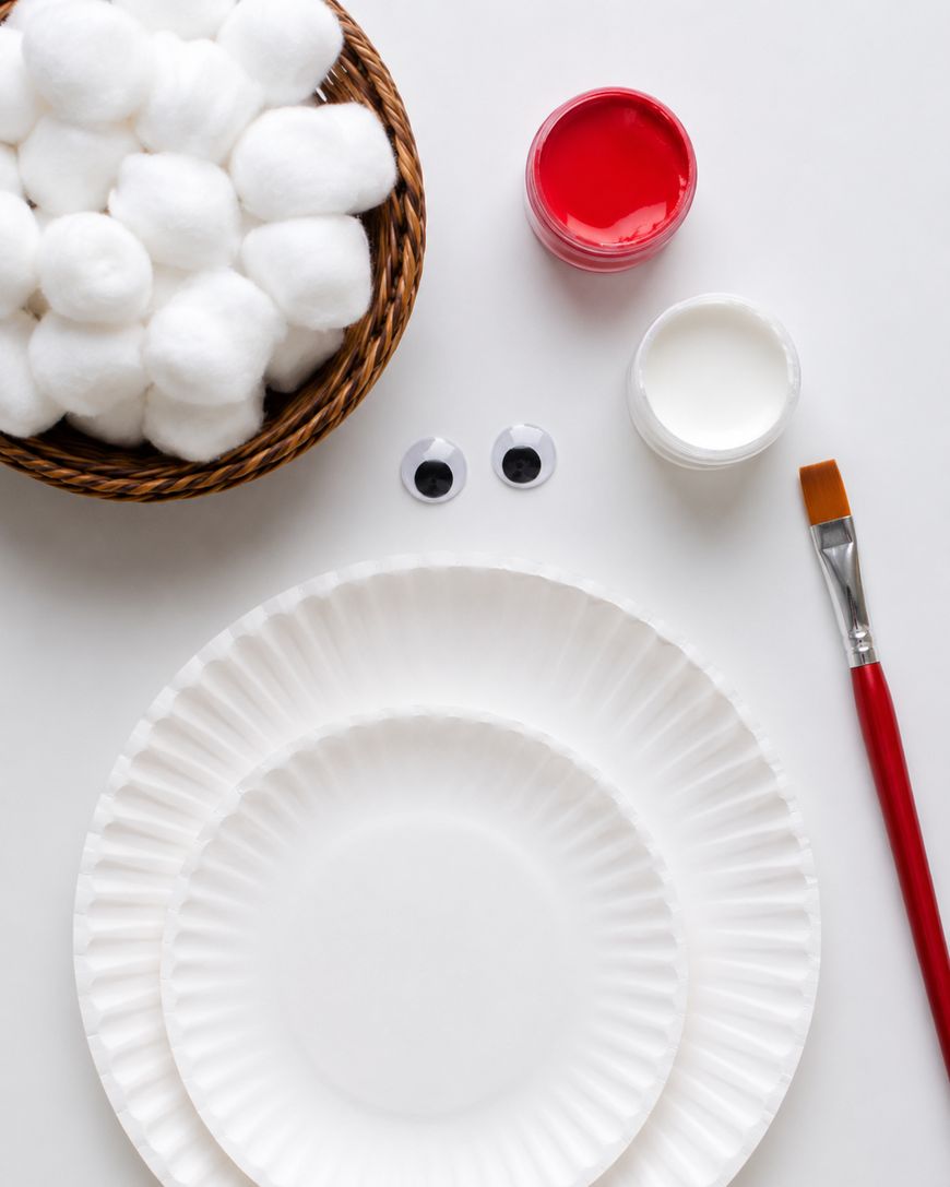A basket of cotton balls, two paper plates, googly eyes, red and white paint jars, and a paintbrush arranged on a white surface.