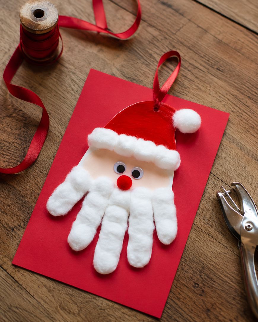 A handmade Santa card features a handprint with cotton balls for the beard and hat trim, googly eyes, and a red nose, placed on a red paper background with ribbon, a spool of thread, and a hole punch nearby.