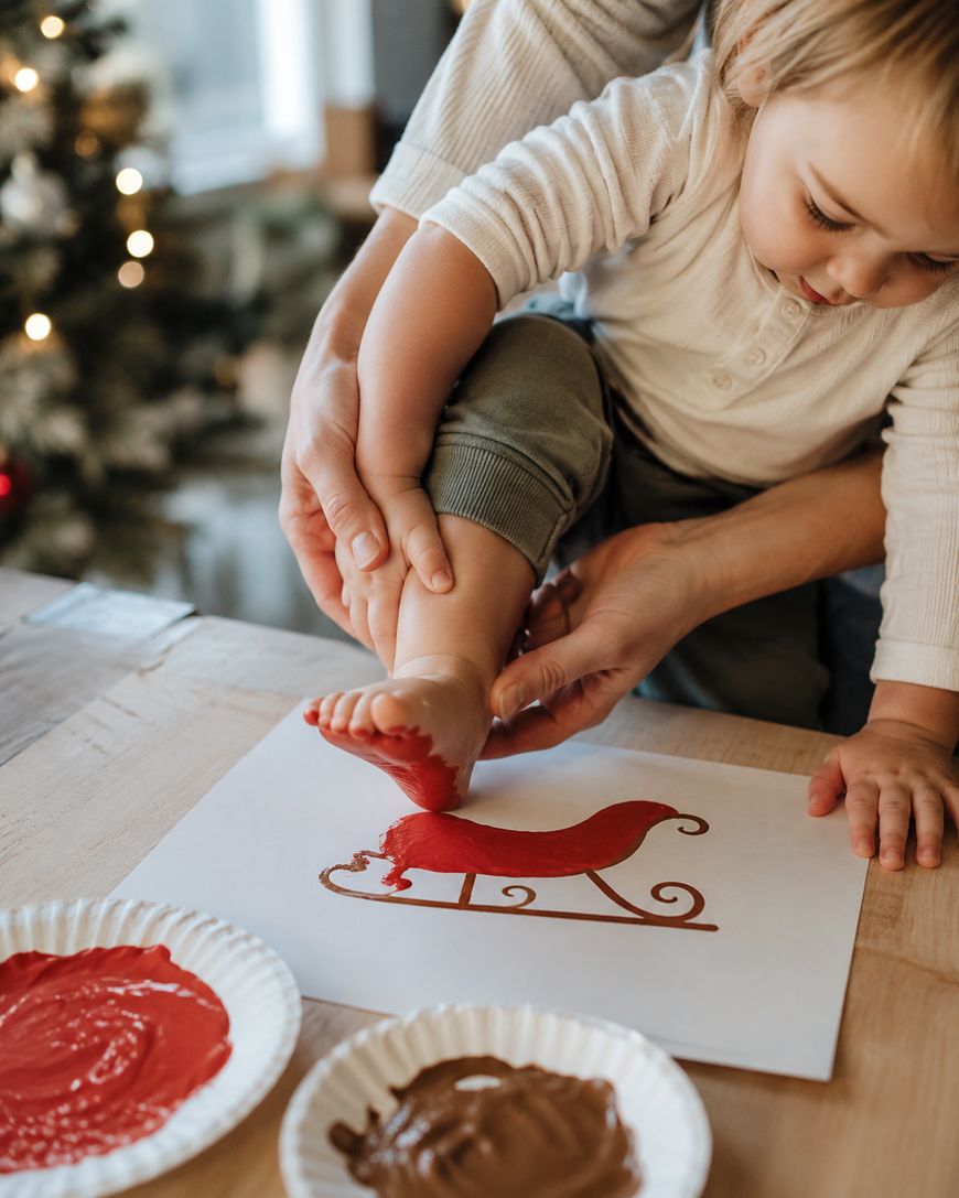 An adult helps a child press their red-painted foot onto paper with a red sleigh outline. Paint plates sit on the table. A Christmas tree is blurred in the background.