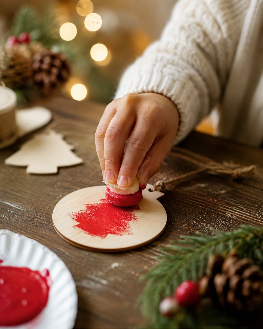 A person paints a wooden ornament with red paint using a small sponge. Christmas decorations, pine branches, and twine are on the table, with soft festive lights in the background.