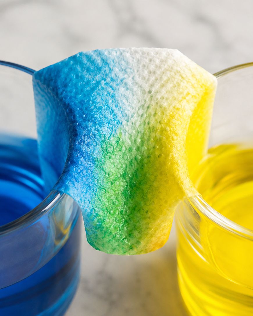 A paper towel bridges two glasses, one with blue liquid and one with yellow liquid, causing the colors to blend into green on the towel.