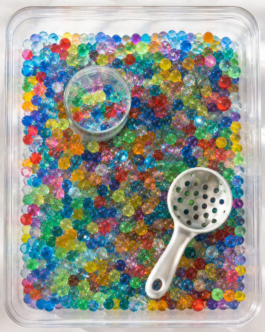 A clear tray filled with colorful water beads, a small clear cup partially filled with beads, and a white plastic scoop with holes, all on a light background.