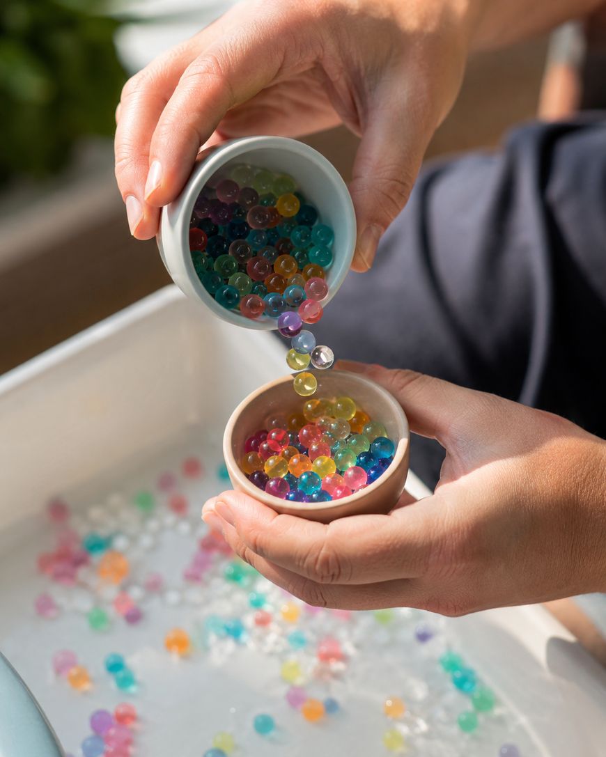 Two hands holding small bowls, with one bowl pouring colorful water beads into the other over a white tray filled with more scattered beads.